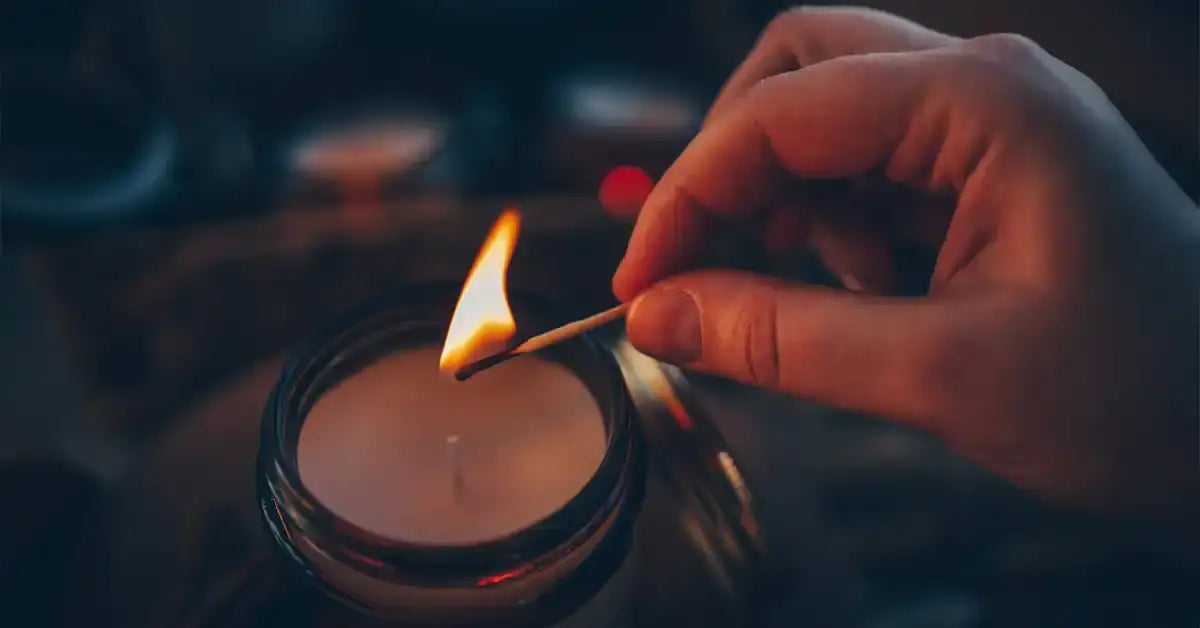 A hand lighting a natural soy candle with a wooden match in a glass jar.