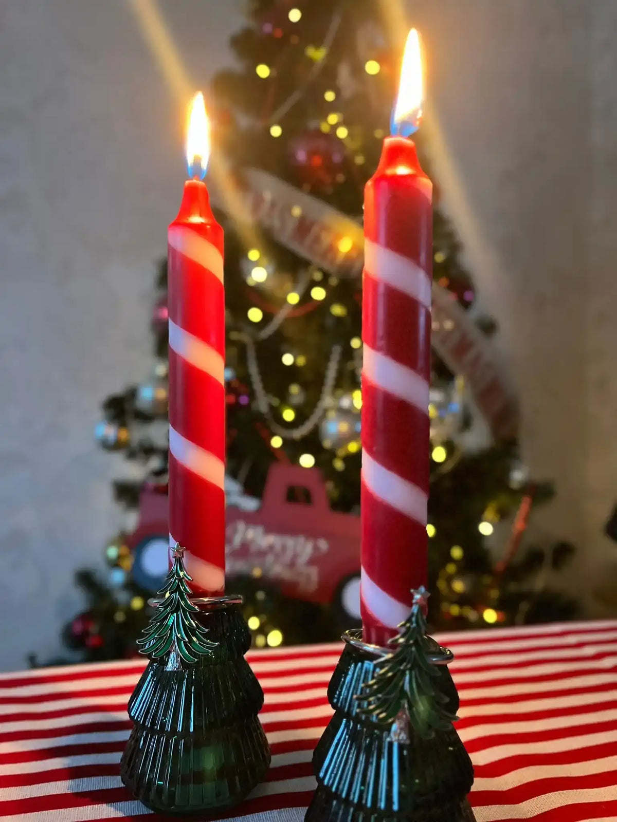 Two lit red and white striped candy cane candles in tree-shaped holders with a blurred Christmas tree.
