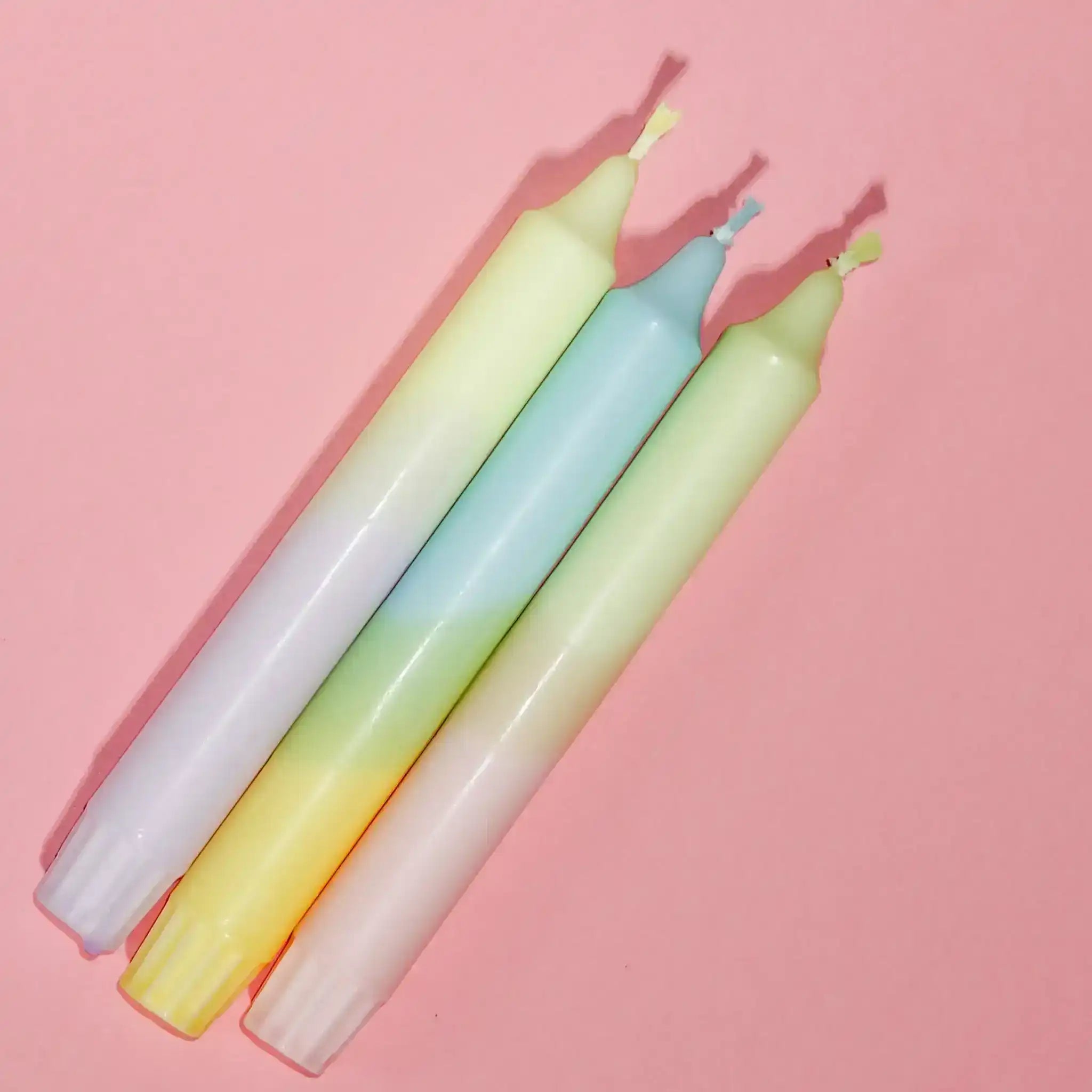 Three pastel rainbow gradient taper candles lying on a pink background