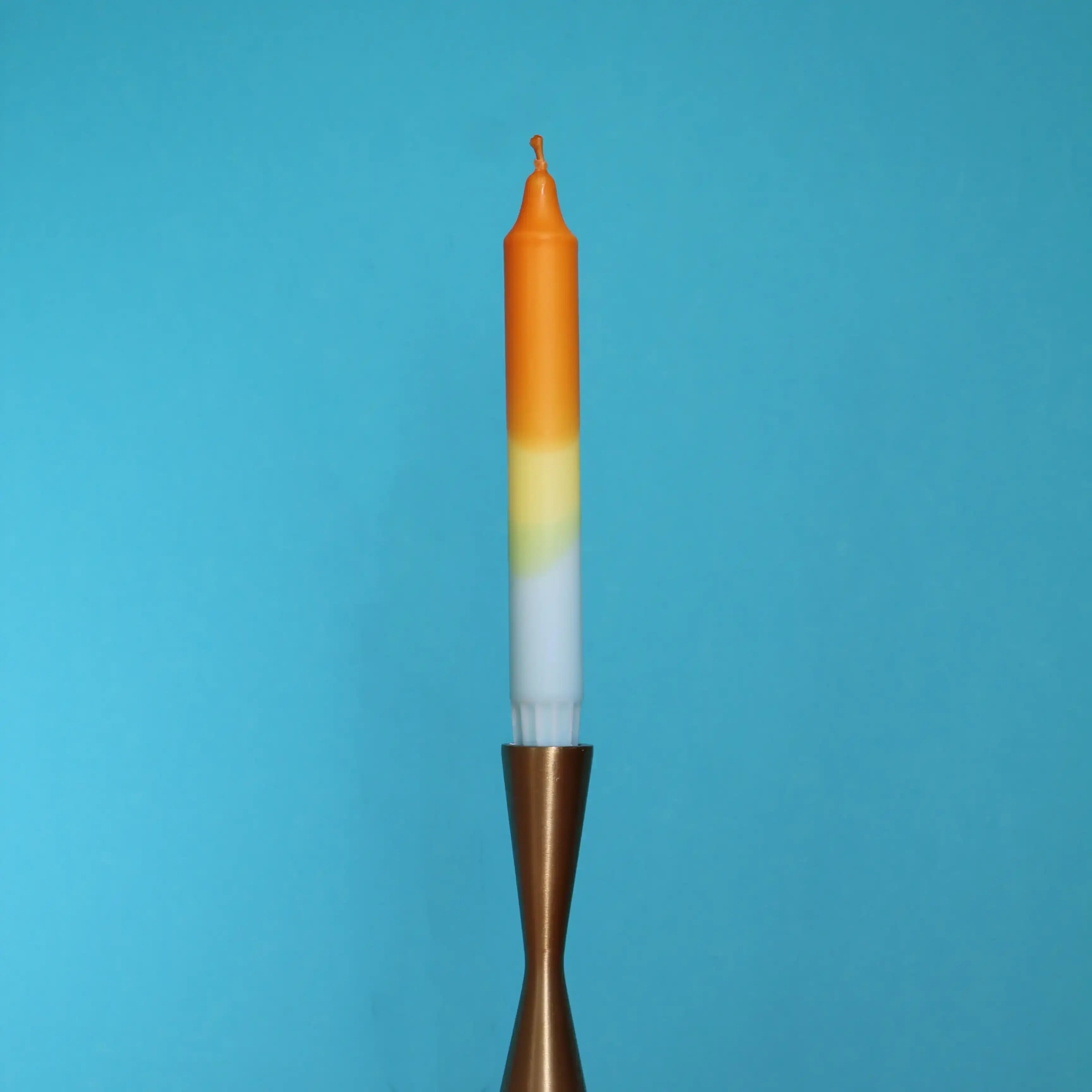 Gradient orange, yellow, and white taper candle in a brass holder against a blue background.