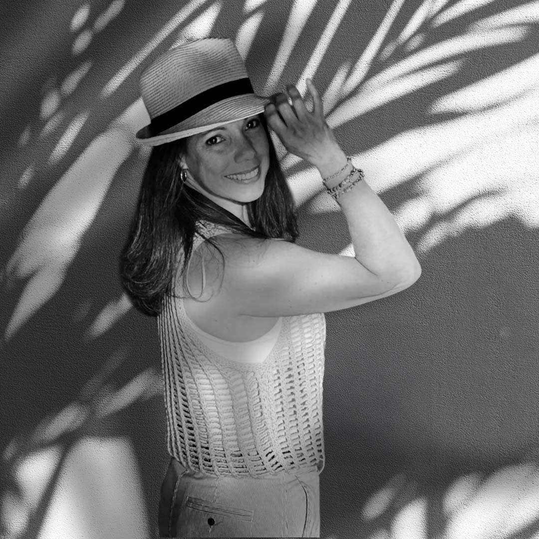 Smiling woman in a sun hat and crochet top standing against a wall with palm leaf shadows.