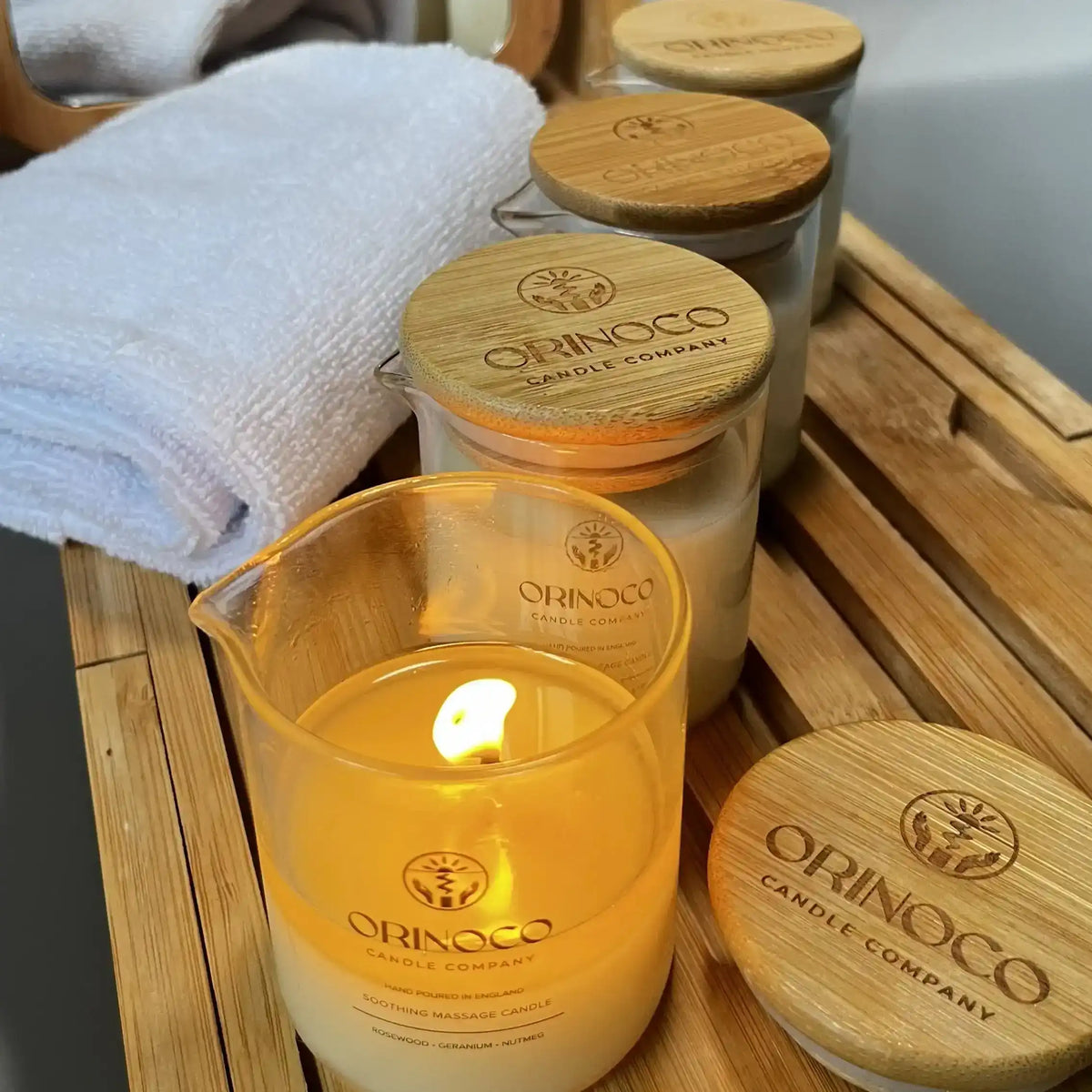 Lit Orinoco Skincare soothing massage candle on a wooden bath tray with white towels.