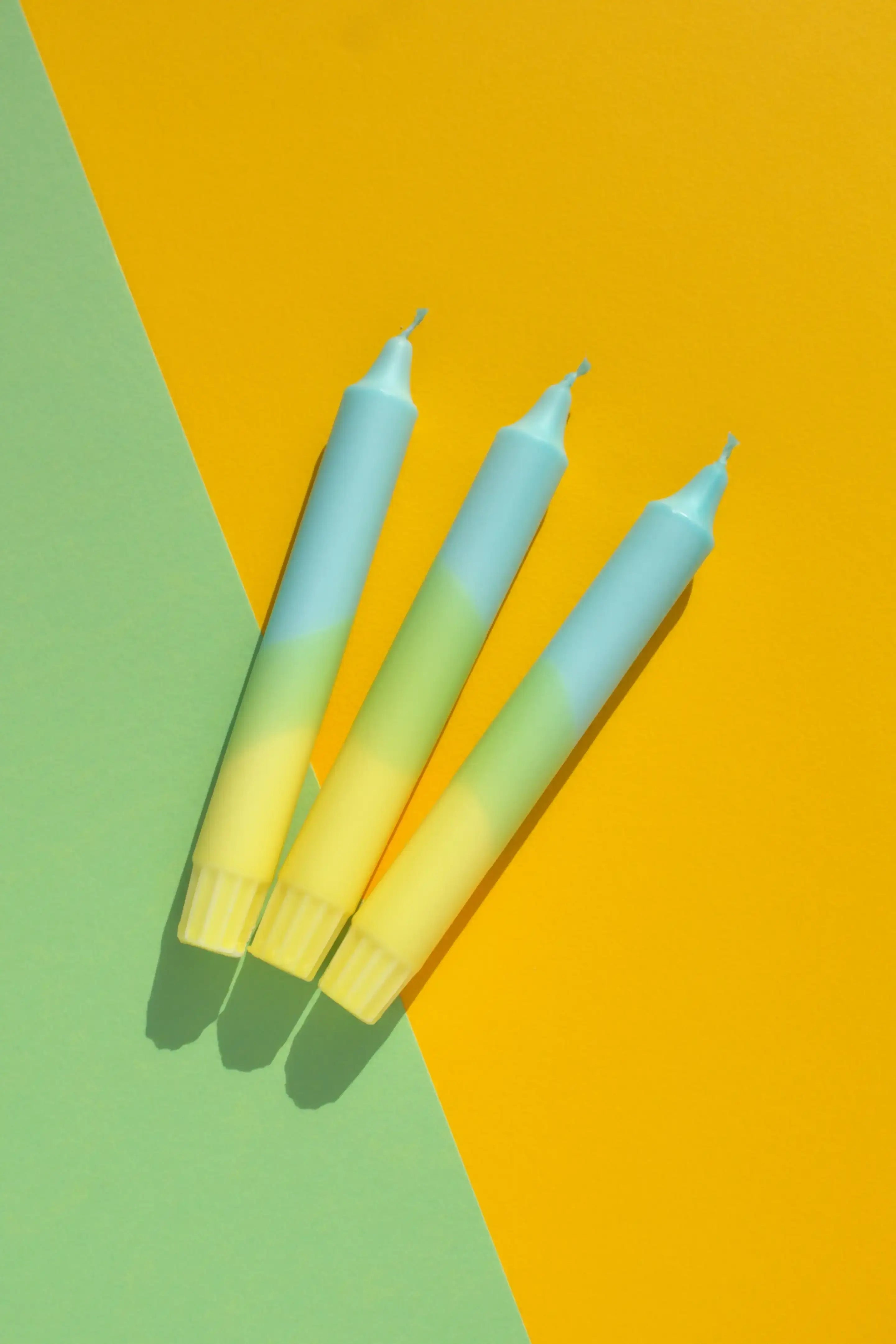 Three gradient blue, green, and yellow taper candles on a split green and yellow background.