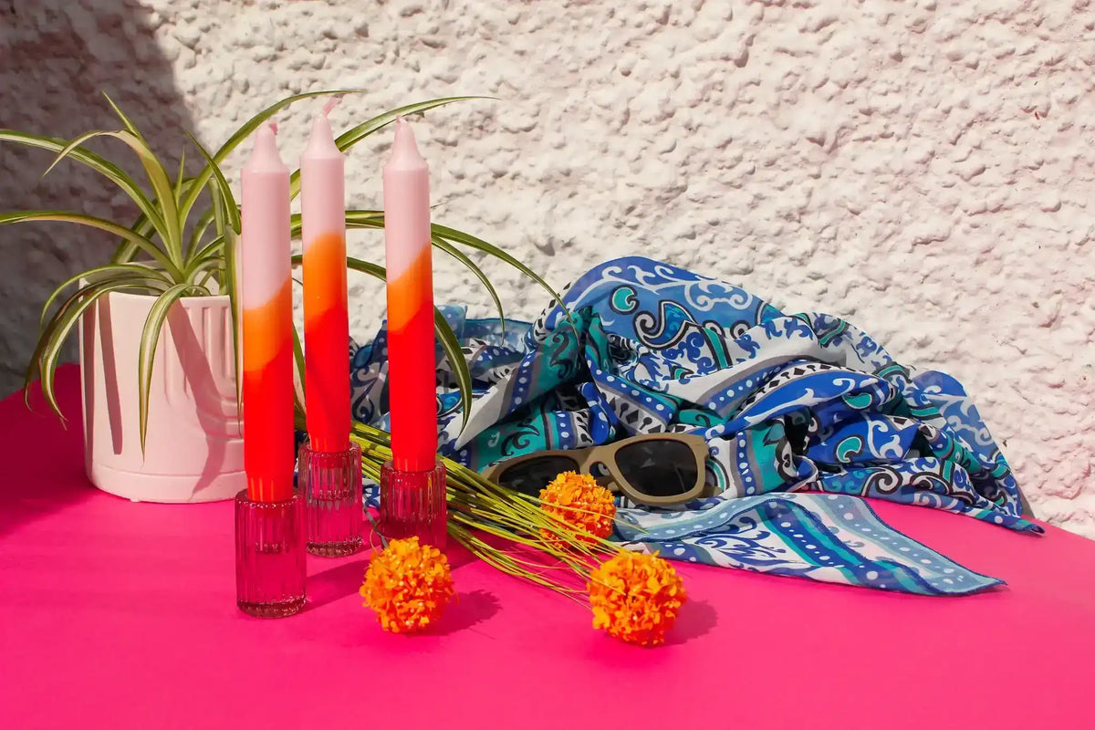 Still life with pink and orange gradient candles, blue patterned scarf, and orange flowers.