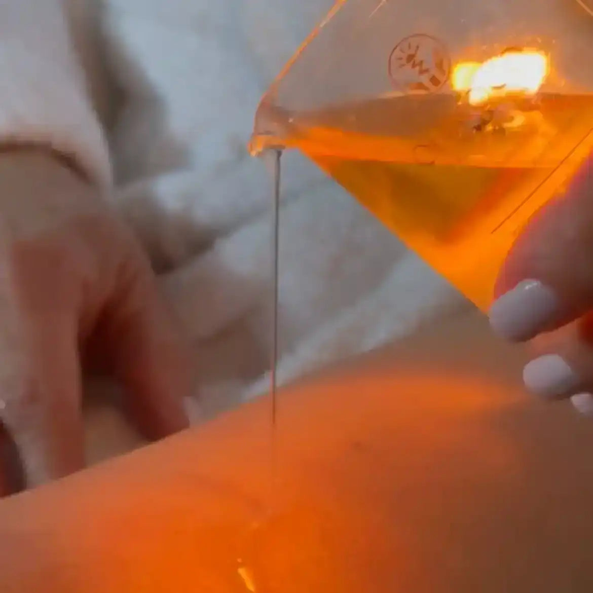 Golden body oil being poured from a glass beaker onto smooth skin for a spa-like treatment.