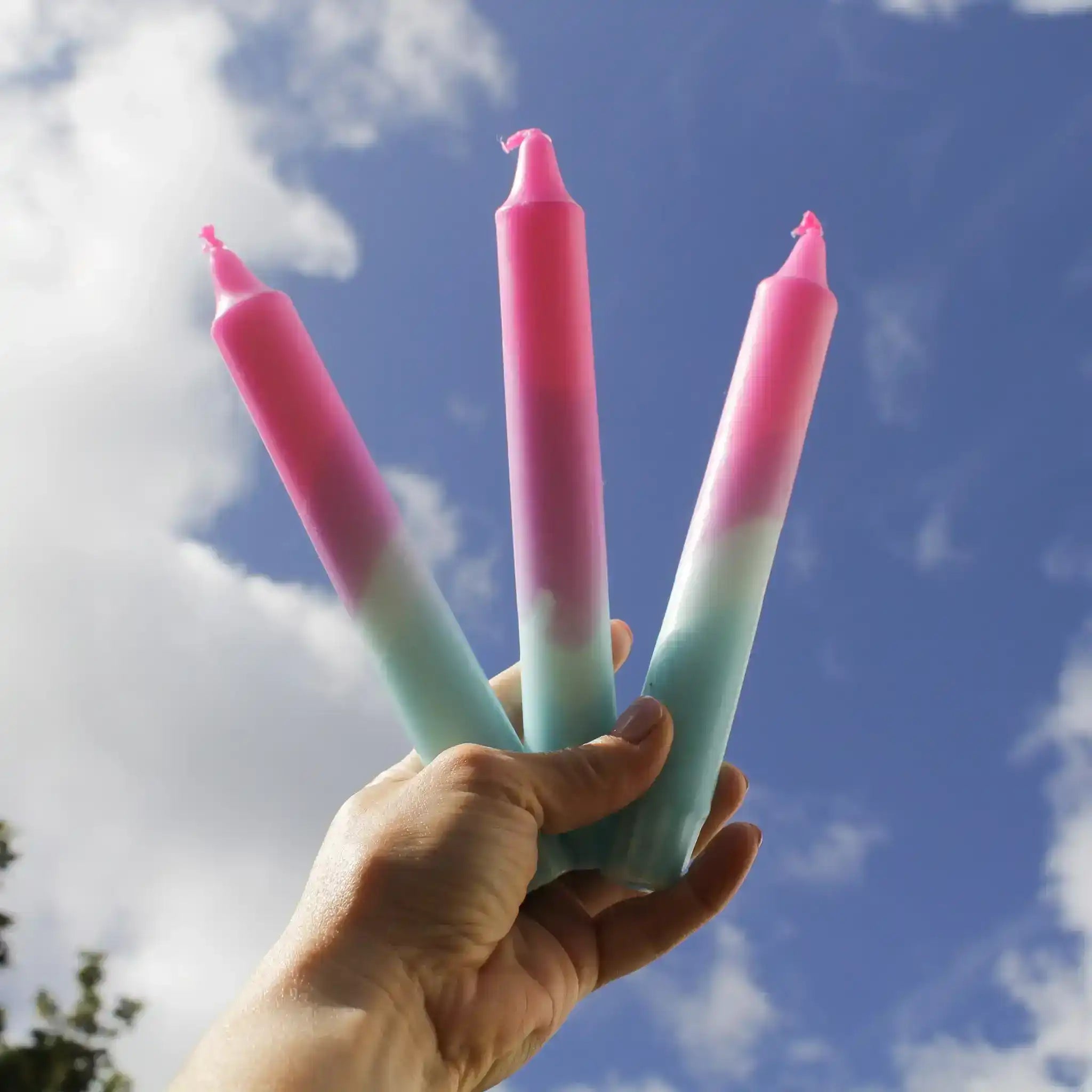A hand holding three pink and blue gradient taper candles against a blue sky with clouds.