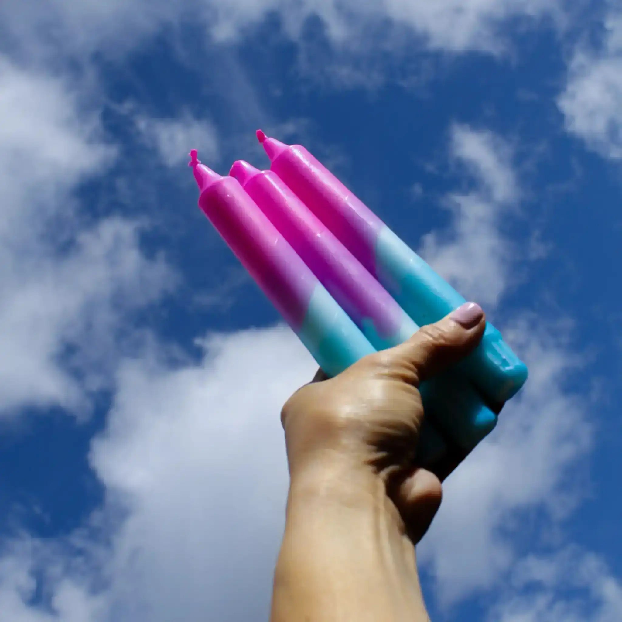Hand holding a bundle of pink and blue gradient tapered candles against a blue cloudy sky