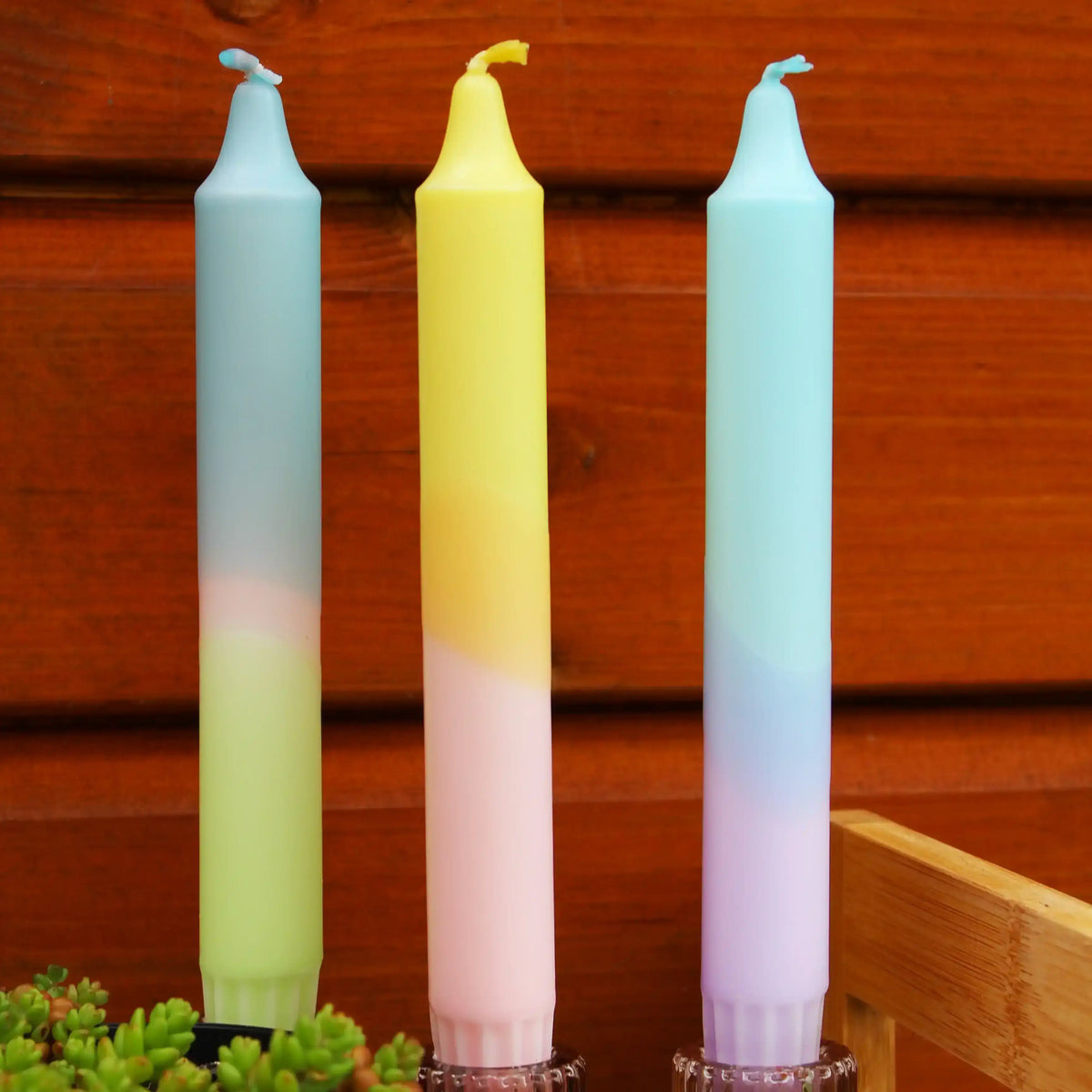 Three pastel gradient taper candles in blue, yellow, and pink against a wooden background.