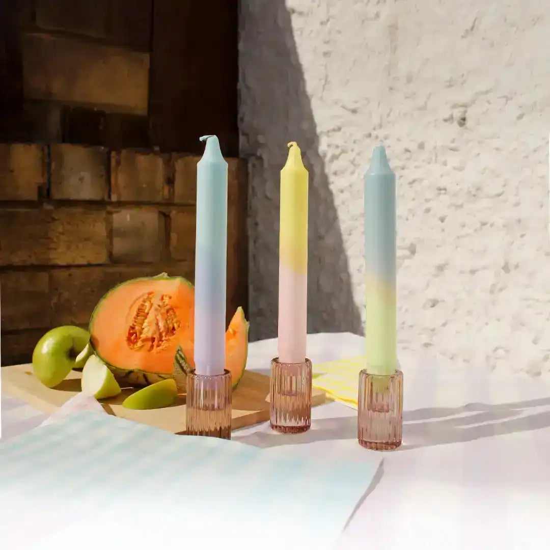 Three colorful gradient taper candles in pink glass holders next to a sliced melon.