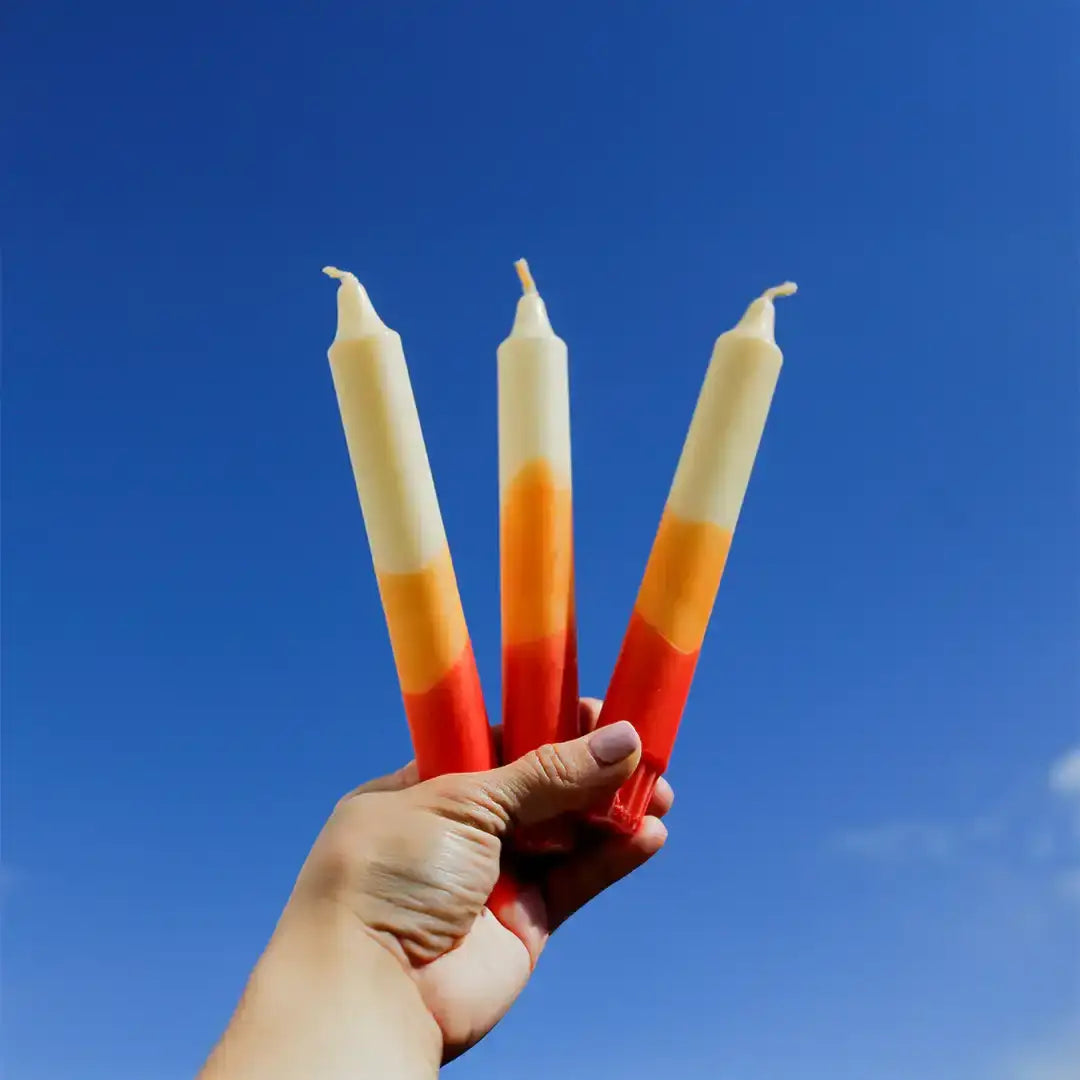 Hand holding three sunset-colored gradient taper candles against a clear blue sky.