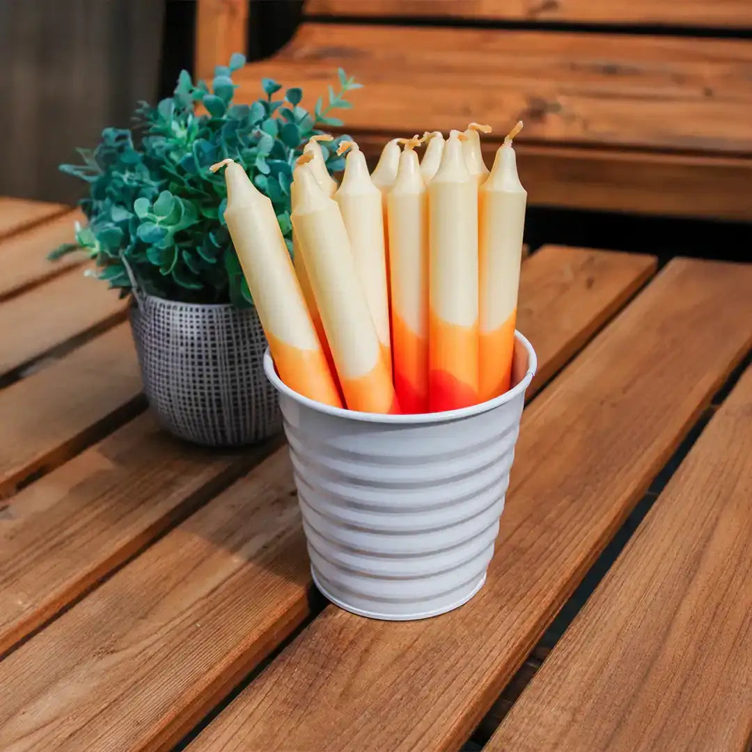 Assorted cream and orange tapered candles in a white ribbed metal bucket on a wooden table.