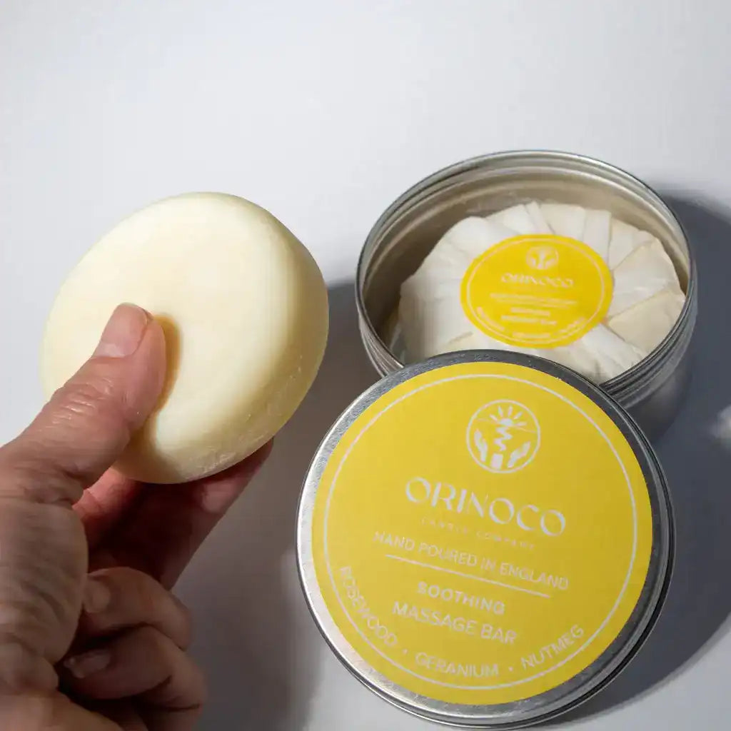 A hand holding a white Orinoco Soothing Massage Bar next to its yellow tin container.