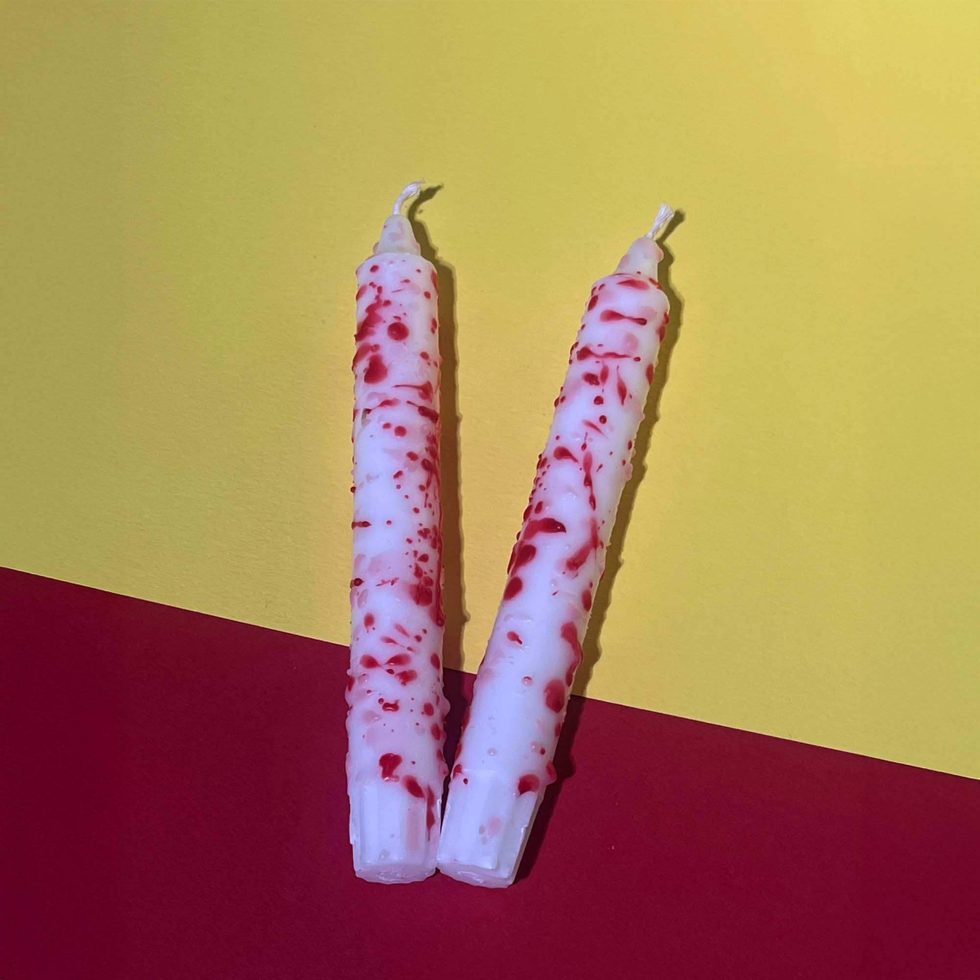 Two white taper candles with red speckled patterns on a yellow and red background.