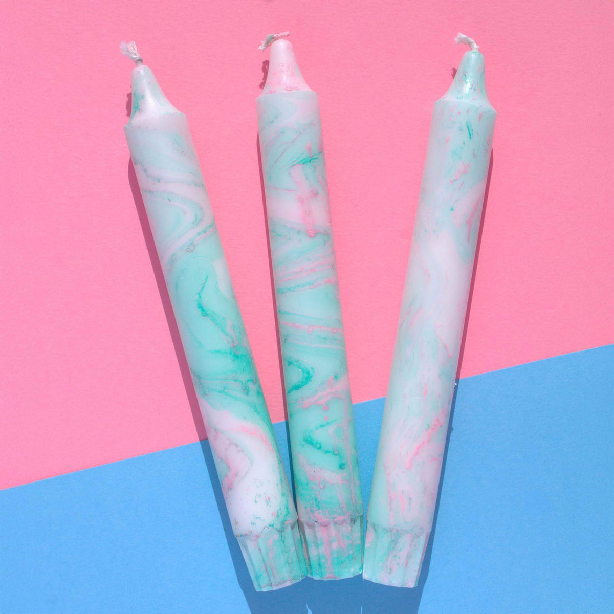 Three tall marbled candles in pastel pink and mint green on a pink and blue background.