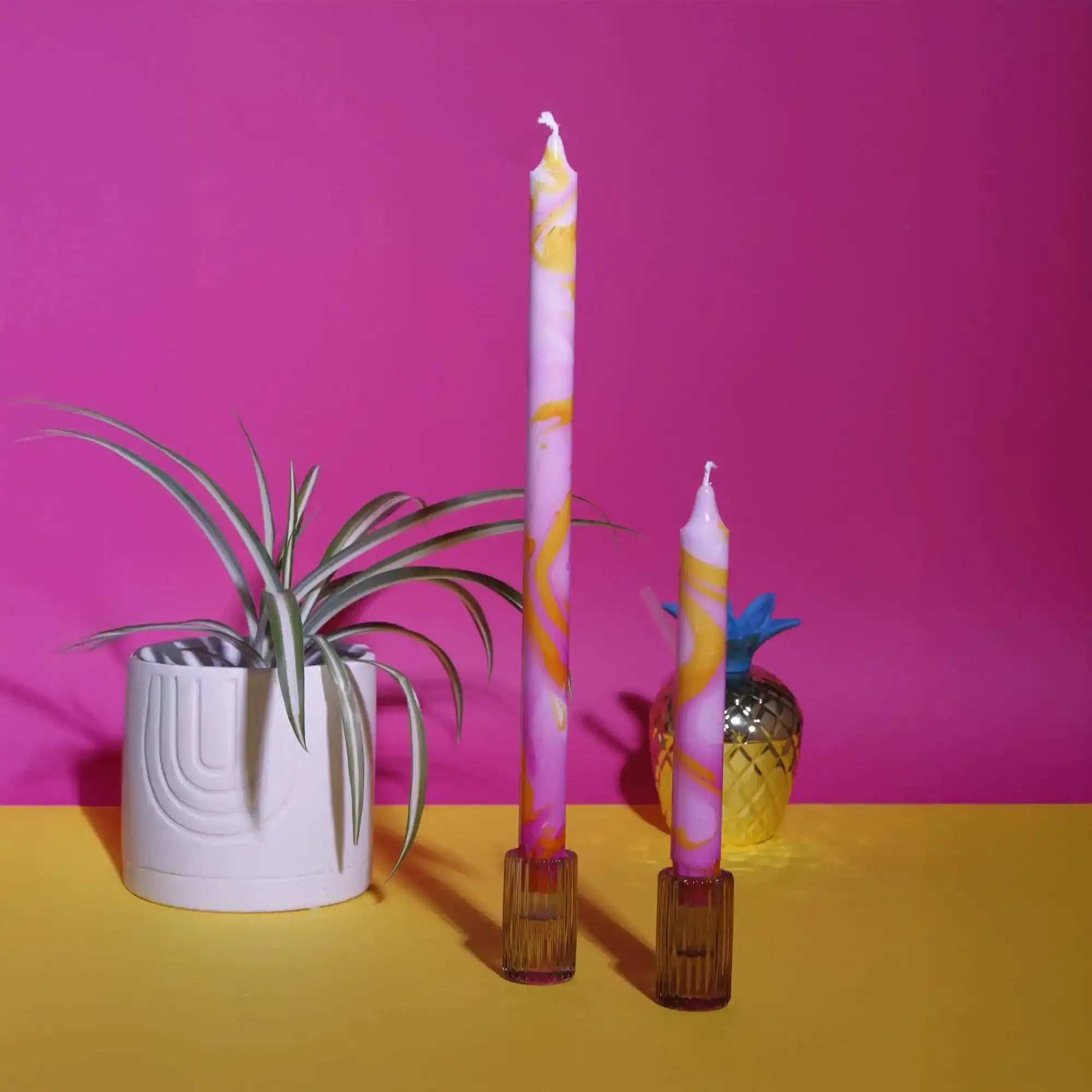 Two tall pink and orange swirl candles in glass holders with a potted plant and gold pineapple.