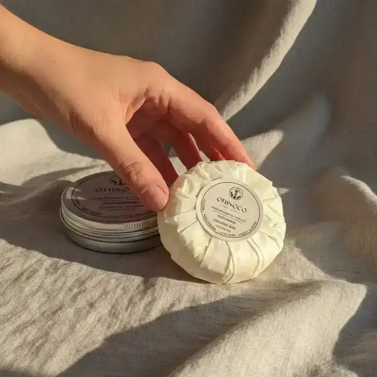 A hand holding an Orinoco skincare product next to its metal tin on a neutral fabric background.